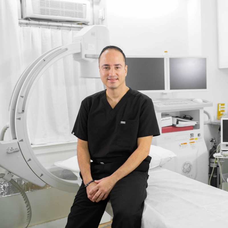 Dr. Montoya positioned in a bright, modern clinic room equipped with advanced medical machinery, including a large C-arm imaging system to his left.